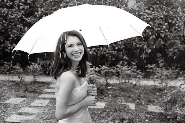 black and white photo - beautiful bride holding standing in the pouring rain holding umbrella and smiling - photo by North Carolina based wedding photographers Cunningham Photo Artists